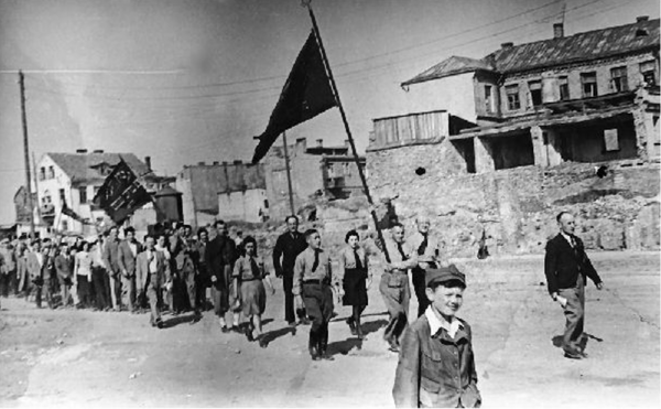A black and white photo in which approximately 30 people are marching down the street. Two flags are being held by members o