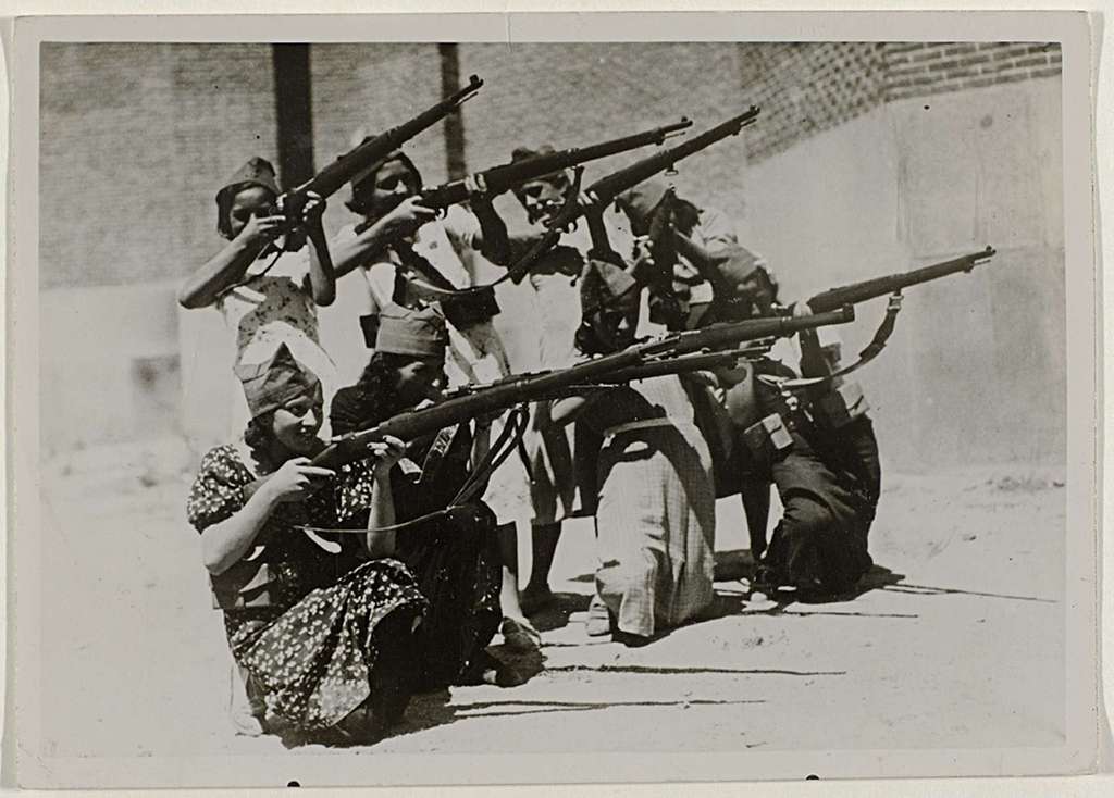 Black and white image of a group of about eight young women wearing dresses pointing rifles toward the right. Description: A republican regiment of women practices with rifles in the streets of Madrid, August 3, 1936 during the Spanish Civil War. Source: Rijksmuseum.