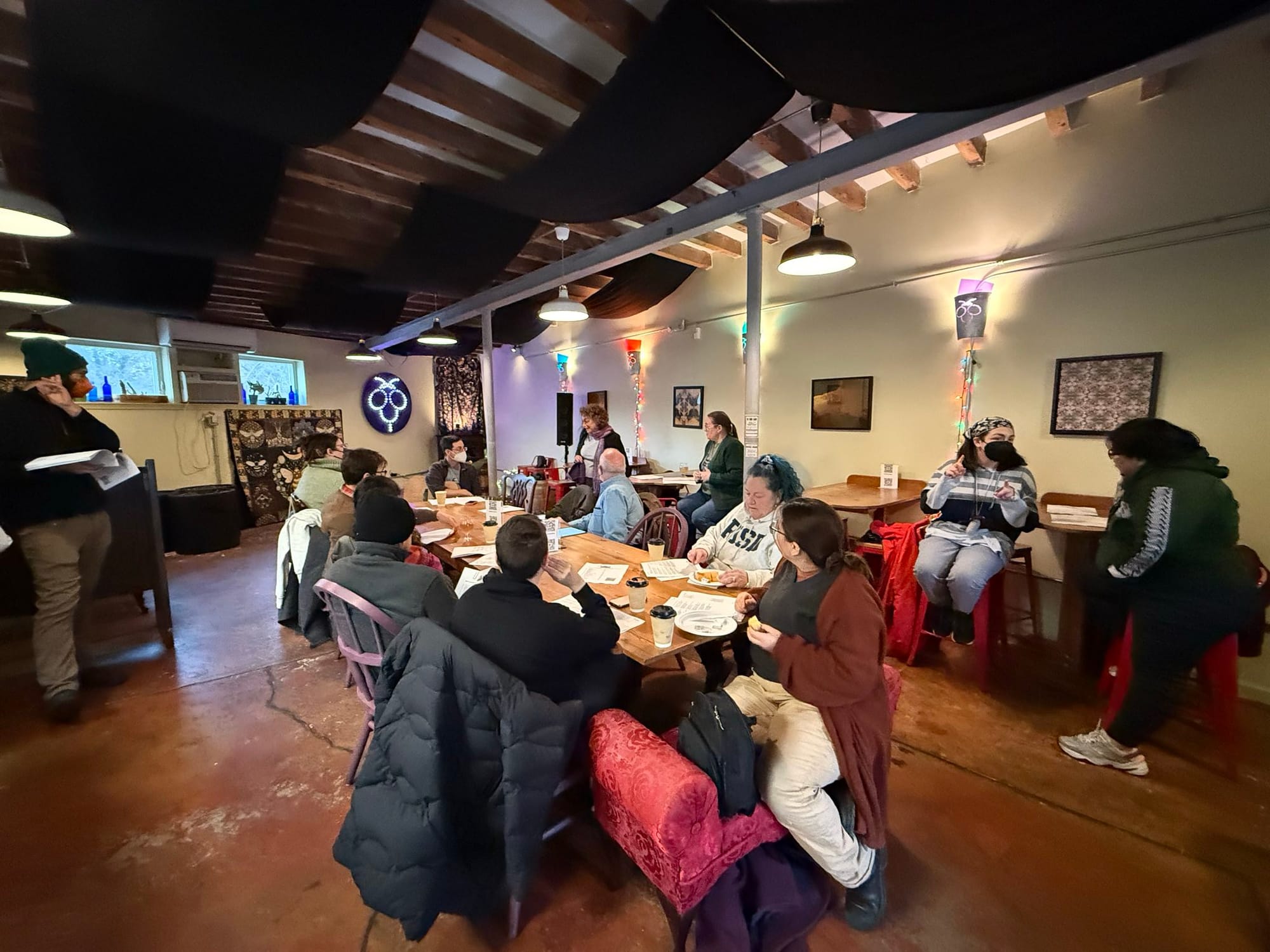 Approximately 13 people are gathered in a small meeting space with wooden tables.