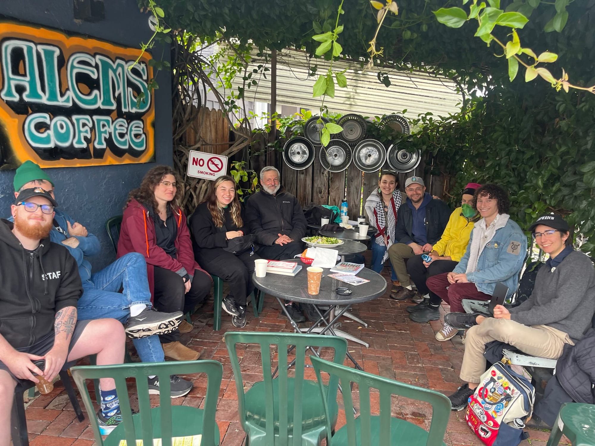 A group of 13 people seated in a semicircle at an outdoor dining area. There is graffiti that reads "Alems Coffee"