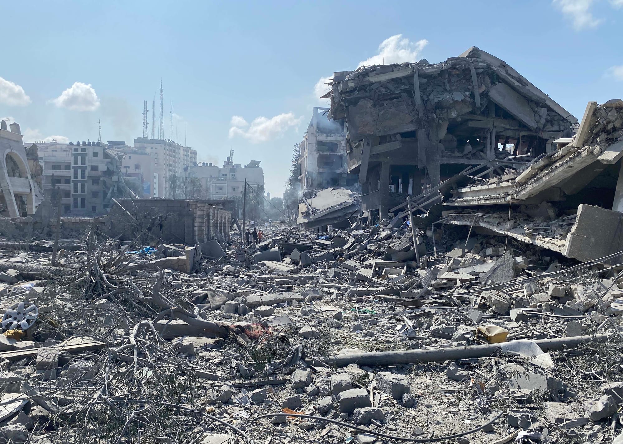 Digital photograph of destroyed buildings in Gaza, the ground covered in debris and rubble, with three people in the background surveying the damage.